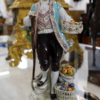 Bei dem Produkt handelt es sich um die Capodimonte-Figur der Königlichen Porzellanmanufaktur Neapel mit dem Titel „Obst- und Blumenhändler“. Sie zeigt eine Person in traditioneller Kleidung, darunter einen schwarzen Hut, einen blauen Mantel und eine gemusterte Weste, die einen Stock und einen Obstkorb hält. Die Figur steht auf einem aufwendig verzierten Sockel mit einem zusätzlichen Obstkorb zu ihren Füßen.