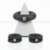 Auf beiden Seiten eines weißen, spitzen Ständers sind zwei schwarze Ringe aus der Kollektion „Set/Ring Kautschuk“ ausgestellt, die jeweils mit einem Diamanten verziert sind. Diese Ringe zeichnen sich durch eine matte Textur und ein schlichtes Design aus, wobei jeder Ring einen einzelnen Diamanten zeigt. Der größere Ring, der vom Ständer gehalten wird, ist ähnlich gestaltet und passt perfekt zur eleganten Ästhetik dieser Kollektion.