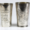 Ausgestellt sind zwei Becher aus poliertem Silber. Der linke Becher ist mit eingravierten Signaturen verziert, während der rechte ein kompliziertes Drachendesign mit Wolkenmotiven und Inschriften im asiatischen Stil zeigt. Beide haben eine reflektierende Oberfläche.