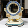 Eine Keramikskulptur mit dem Titel „Karlsruhe Majolika Keramik Manufaktur – Vier Putti – Modell Nr. 1060“, entworfen von Wilhelm Kollmar, zeigt vier Engelchen, die knien und eine große, runde Struktur stützen, die mit blauen Blumen verziert ist. Diese Engelchen stehen auf einem sechseckigen Sockel, haben goldenes Haar und gelbe Schärpen, im Hintergrund sind Kunstwerke und Rahmen zu sehen. Das Stück enthält kein Uhrwerk.