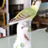 Eine Grünspechtfigur der Meissen Porzellan Manufaktur mit grünem und gelbem Gefieder thront auf einem kunstvoll gestalteten weißen Sockel mit kleinen Blumenmotiven. Sie wird auf einer Glasoberfläche präsentiert, im Hintergrund sind die Kunstwerke sanft verschwommen.
