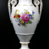 Die Amphoren-Vase der Meissen Porzellan Manufaktur mit einem Blumendekor aus rosa, roten, gelben und violetten Blumen, akzentuiert durch einen Goldrand und verzierte Griffe, ist aus weißem Porzellan mit glänzender Oberfläche gefertigt. Sie steht auf einem quadratischen Sockel vor schwarzem Hintergrund.
