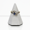 Auf einem grauen, pyramidenförmigen Ständer ist der 585/Bicolor Ring mit Brillant ca. 0,2ct. und Saphir sowie acht Diamantrosen ausgestellt, dessen Goldband mit zwei markanten Edelsteinen verziert ist: einem blauen Saphir und einem klaren Brillanten. Der Ring enthält außerdem mehrere kleinere klare Diamantrosen entlang des Bandes, die auf einem weißen Hintergrund angeordnet sind.
