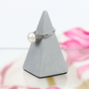 Der antike Ring aus Pt/950 mit Perle und Diamantsplittern wird auf einem grauen, pyramidenförmigen Ständer präsentiert, während der Hintergrund eine unscharfe Mischung aus rosa und weißen Blumenmustern zeigt.