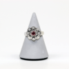 Der 585/Weißgold-Ring, der mit einem leuchtenden Rubin in der Mitte geschmückt und von acht schillernden Diamanten mit insgesamt etwa 0,25 Karat umgeben ist, wird elegant auf einem schlanken, grauen Kegelständer vor einem schlichten weißen Hintergrund präsentiert.