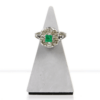 Ein 585er Weißgoldring zeigt in der Mitte einen quadratischen grünen Smaragd, der von einem Kranz aus acht Diamanten im Altschliff umgeben ist. Der Ring wird auf einem dreieckigen grauen Ständer vor weißem Hintergrund präsentiert.