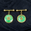 Ein Paar 585/Manschettenknöpfe Gelbgold mit Jade zeigt runde grüne Jadesteine, jeweils akzentuiert mit einem einzigartigen Emblem – einer mit einem Monogramm und der andere mit einem Blumenmuster – vor einem dunklen, strukturierten Stoffhintergrund.