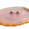 Auf einer rosa-beigen Achatscheibe ist ein Paar Ohrringe mit der Bezeichnung „750/Gelbgold-Ohrstecker mit Diamanten und Amethyst“ angebracht, die aus Goldfassungen mit ovalen violetten Amethysten bestehen, die von kleinen klaren Diamanten umgeben sind.