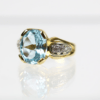 Eine Nahaufnahme des 750/Gelbgold-Rings, in dessen Mitte ein großer Spinell-Edelstein im Ovalschliff zu sehen ist. Das Band ist auf jeder Seite mit kleinen Brillanten verziert und der Ring liegt auf einer reflektierenden Oberfläche vor einem weißen Hintergrund.