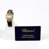 Die Armbanduhr DAU Chopard Happy Diamonds, Referenznummer 20/5523, mit goldenem Gehäuse und braunem Lederarmband, wird auf einem weißen Ständer neben einer dunkelblauen Karte mit der Aufschrift „Chopard-Kundendienst-Garantiezertifikat“ präsentiert.