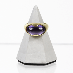 Der 585/Gelbgold-Ring mit Amethyst, der einen großen ovalen violetten Edelstein besitzt, wird auf einem grauen pyramidenförmigen Ständer vor weißem Hintergrund präsentiert.