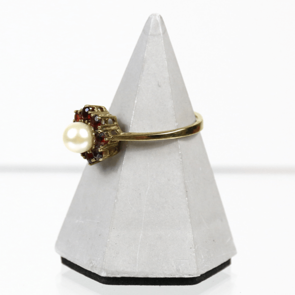 Wir präsentieren den 333/Gelbgold-Ring mit Granat und Perle. Sein Goldband ist mit einer Perle in der Mitte und Akzenten aus rotem Granat verziert und wird elegant auf einem geometrischen, pyramidenförmigen Ständer in Grau präsentiert.