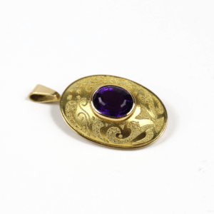 Der Anhänger und die Brosche mit Amethyst aus 750/Gelbgold ist ein ovaler Goldanhänger mit einem kunstvoll gravierten Design und einem markanten dunkelvioletten Amethyst in der Mitte, komplett mit einer kleinen Öse zum Befestigen an einer Kette.