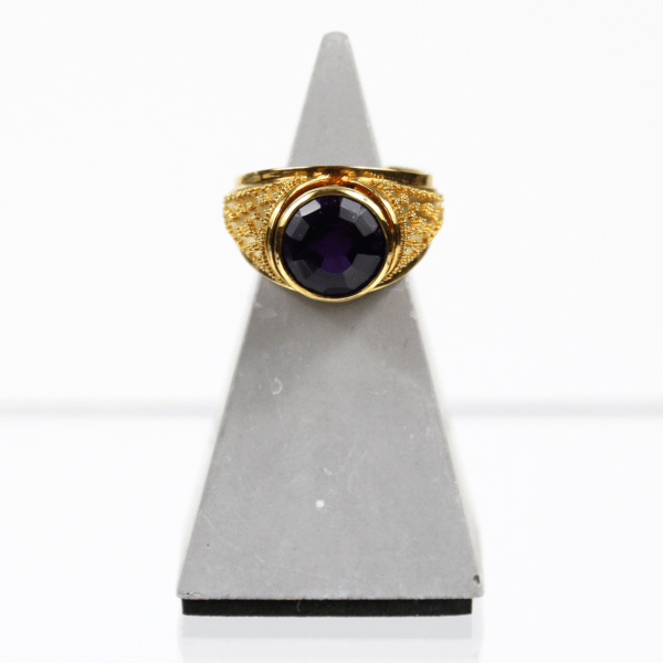 Der 750/Gelbgold-Ring mit Amethyst, der einen großen, runden, dunkelvioletten Edelstein und aufwendige Details auf der Ringschiene aufweist, wird auf einem grauen dreieckigen Ständer vor weißem Hintergrund präsentiert.